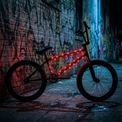 A BMX-style bicycle with glowing red LED light strips along the frame, parked against a graffiti-covered brick wall at night on a wet urban street.