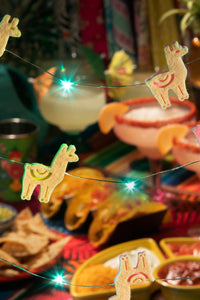 Decorative string lights with llama shapes over a festive table with food and drinks.