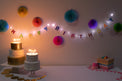 Birthday cake with lit candles, colorful 'Happy Birthday' LED light banner, and decorative lights on a white wall.