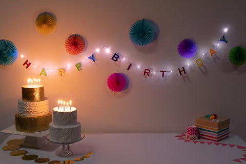 Birthday cake with lit candles, colorful 'Happy Birthday' LED light banner, and decorative lights on a white wall.