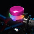 A close-up of a bicycle handlebar bell with a bright pink top and glowing multicolor LED light, mounted on a dark bicycle frame.