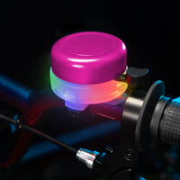 A close-up of a bicycle handlebar bell with a bright pink top and glowing multicolor LED light, mounted on a dark bicycle frame.