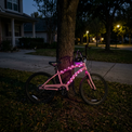 A pink bicycle with glowing pink LED light strips along the frame, parked against a tree on a grassy lawn at dusk in a suburban neighborhood.