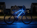 A bicycle at night with bright blue LED light strips along the frame, parked on a residential street with houses blurred in the background.