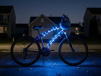 A bicycle at night with bright blue LED light strips along the frame, parked on a residential street with houses blurred in the background.