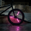 A black BMX bicycle wheel with glowing pink LED light bars on the spokes, photographed in a dark skatepark environment.